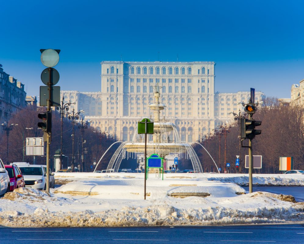 The large Parliament Building in Bucharest city in winter. There is a blue sky and snow on the ground.