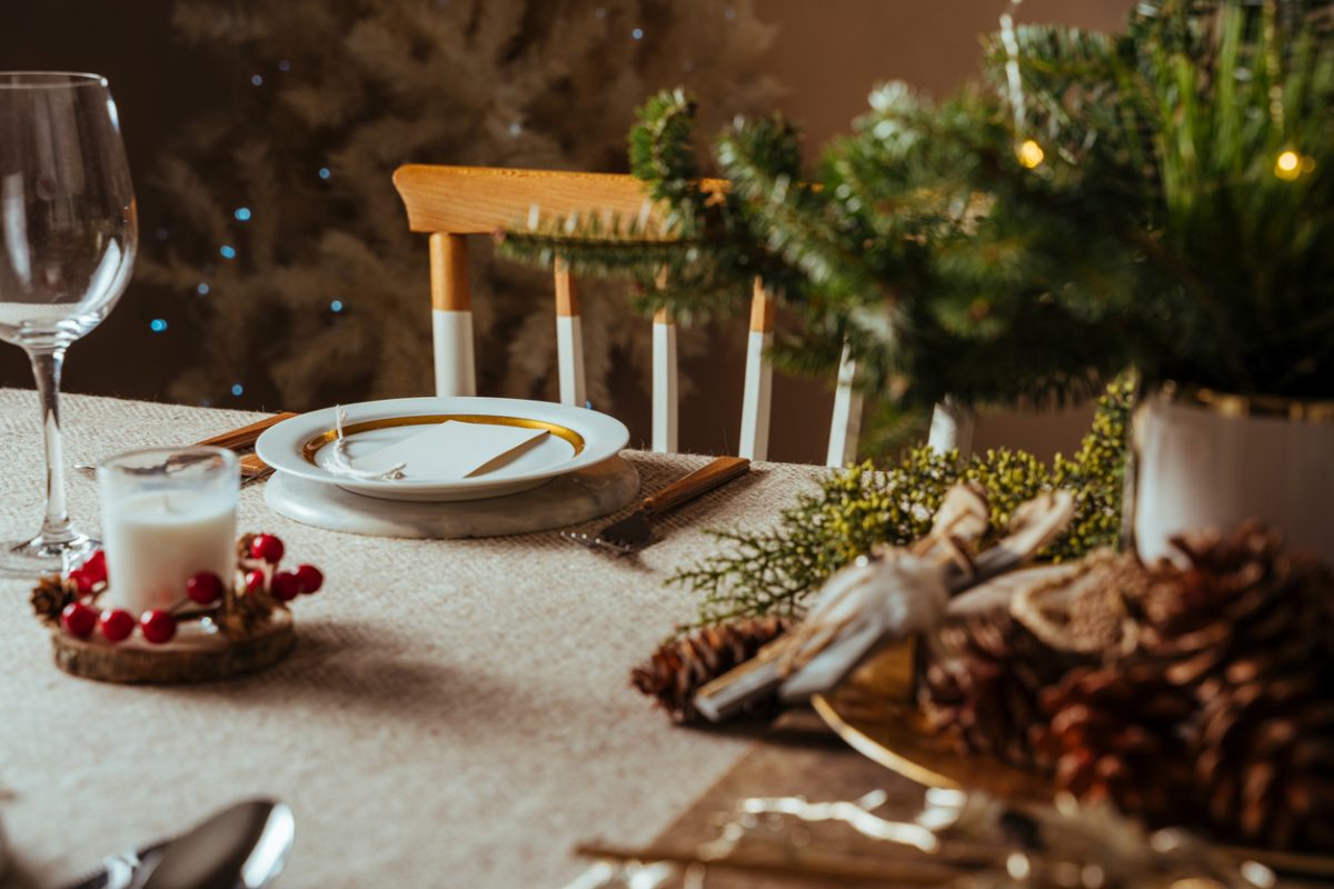 Homely christmas table setting, decorated with pine branches , pine cones, and rustic tablecloth surrounded by Nordic decoration and lights, with the illuminated tree in the background. Family dinner.