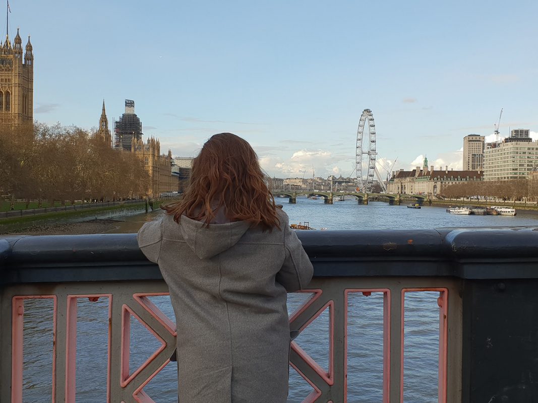 Girl looking over the River Thames and London Eye from a bridge. She is wearing a grey coat. The sky is blue. 