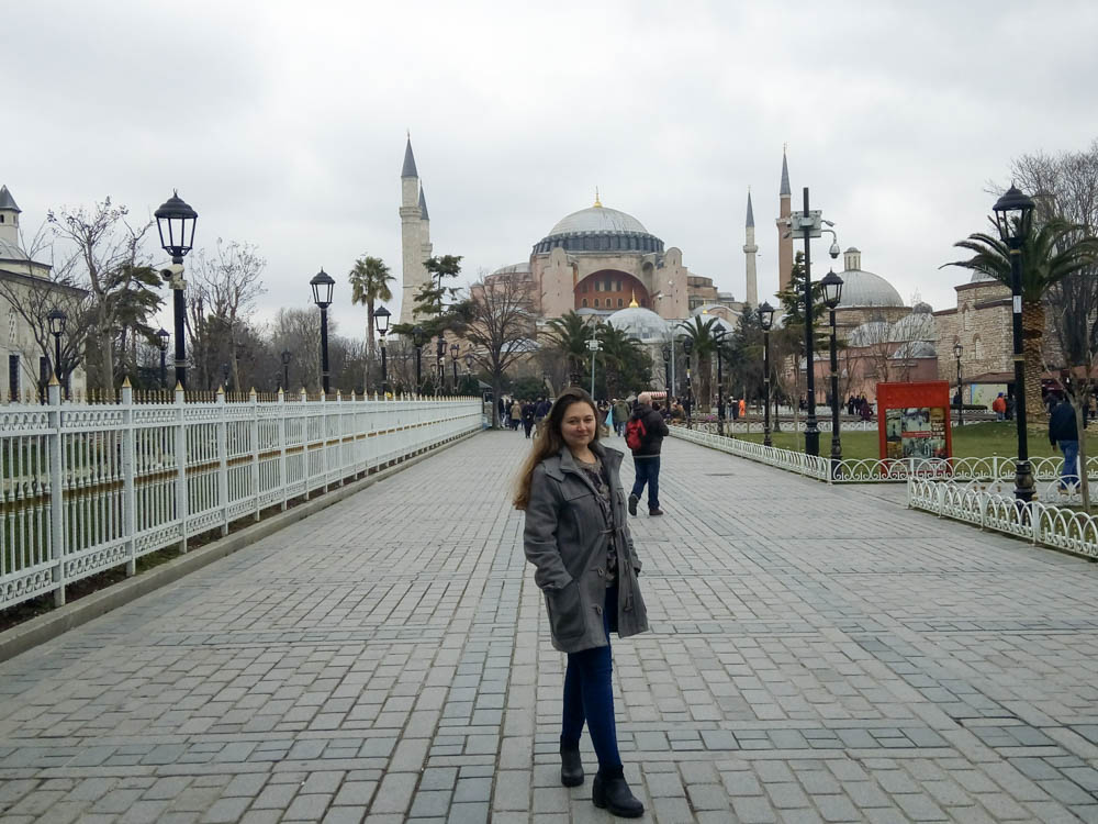 Girl standing infront of Hagia Sophia in Istanbul. The sky is grey and she is wearing trousers and a coat. 