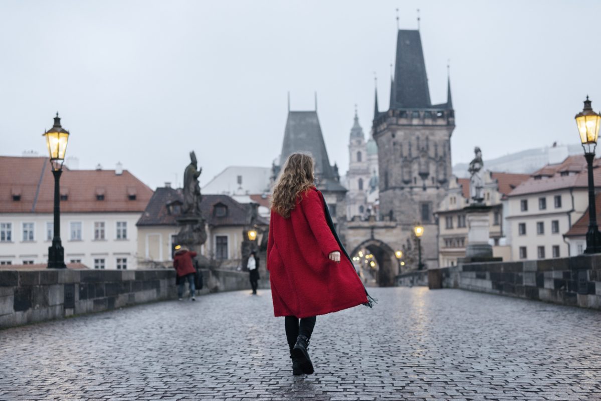 Female tourist wearing red coat walking alone on the street of Prague during the foggy, winter dawn