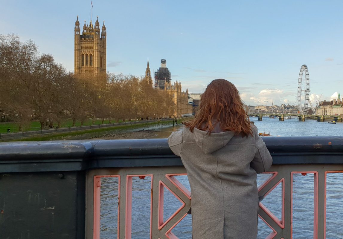 Girl looking out over the River Thames wearing a grey jacket. The London Eye is in the background. 