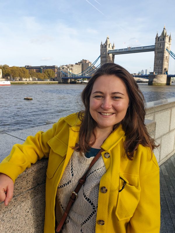 Girl smiling infront of Tower Bridge in London. She's wearing a bright yellow jacket and the River Thames is in the background. 