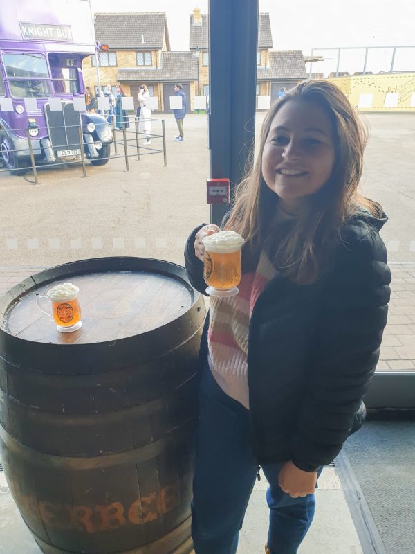 Girl drinking a butterbeer at the Harry Potter Studio tours in London. She is standing next to a barrel. The Knight bus is in the background. 