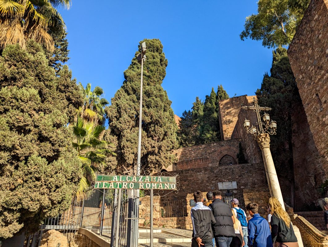 Sign indicating to the Roman Theatre in Malaga. There are trees in the background and the sky is a bright blue.