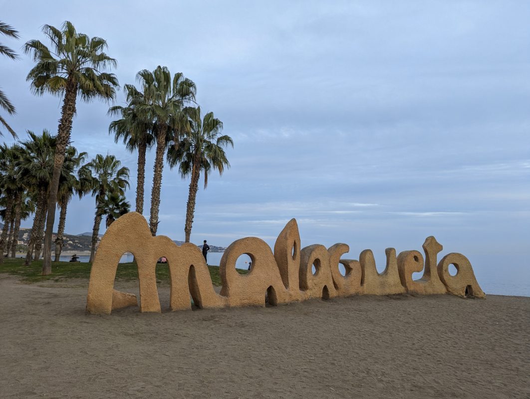 Lettering of Malagueta with golden sand and palm trees in the background. 