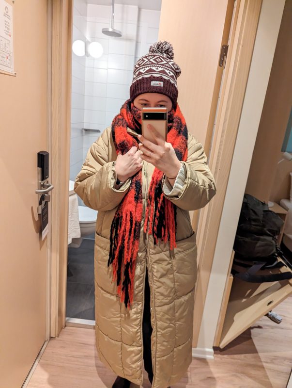Claire is taking a photo in her full-length hotel mirror, wearing a red and black checked scarf, burgundy hat and long beige coat.