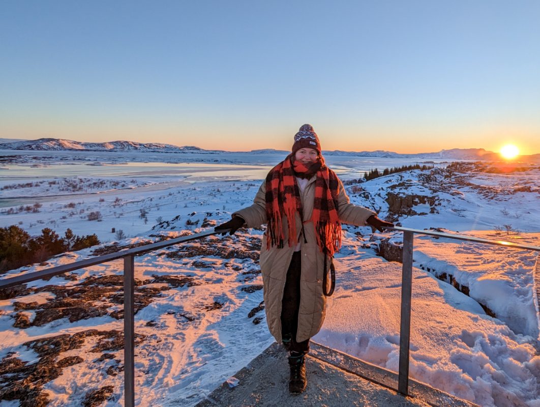 Girl standing on the North American tectonic plate at Thingveller National Park on the Golden Circle in Iceland, wearing a beige coat, red and black checked scarf and burgundy hat, with the view of "no man's land" in the background before the Eurasian plate.