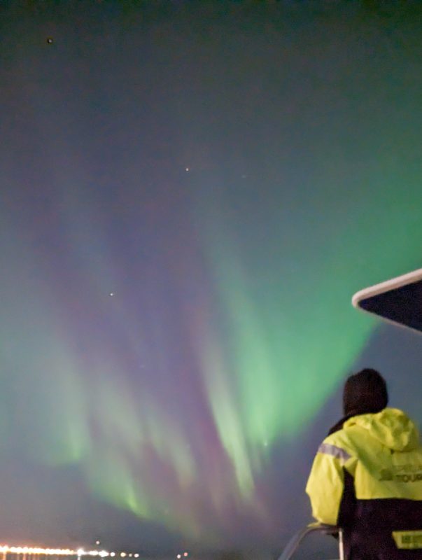 Green flashes of Northern Lights with a man standing with a reflective coat in the foreground.