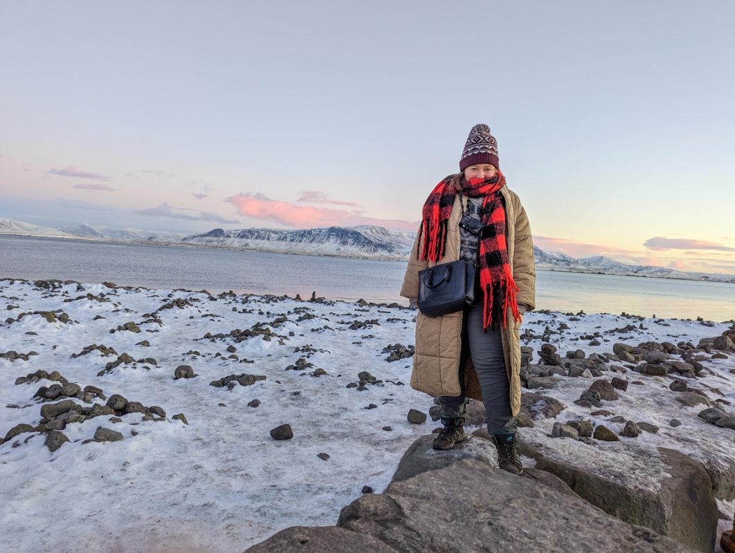 Girl standing infront of the coastline, with mountains in the background. She is wearing a thick, long coat, scarf and gloves in Reykjavik in winter