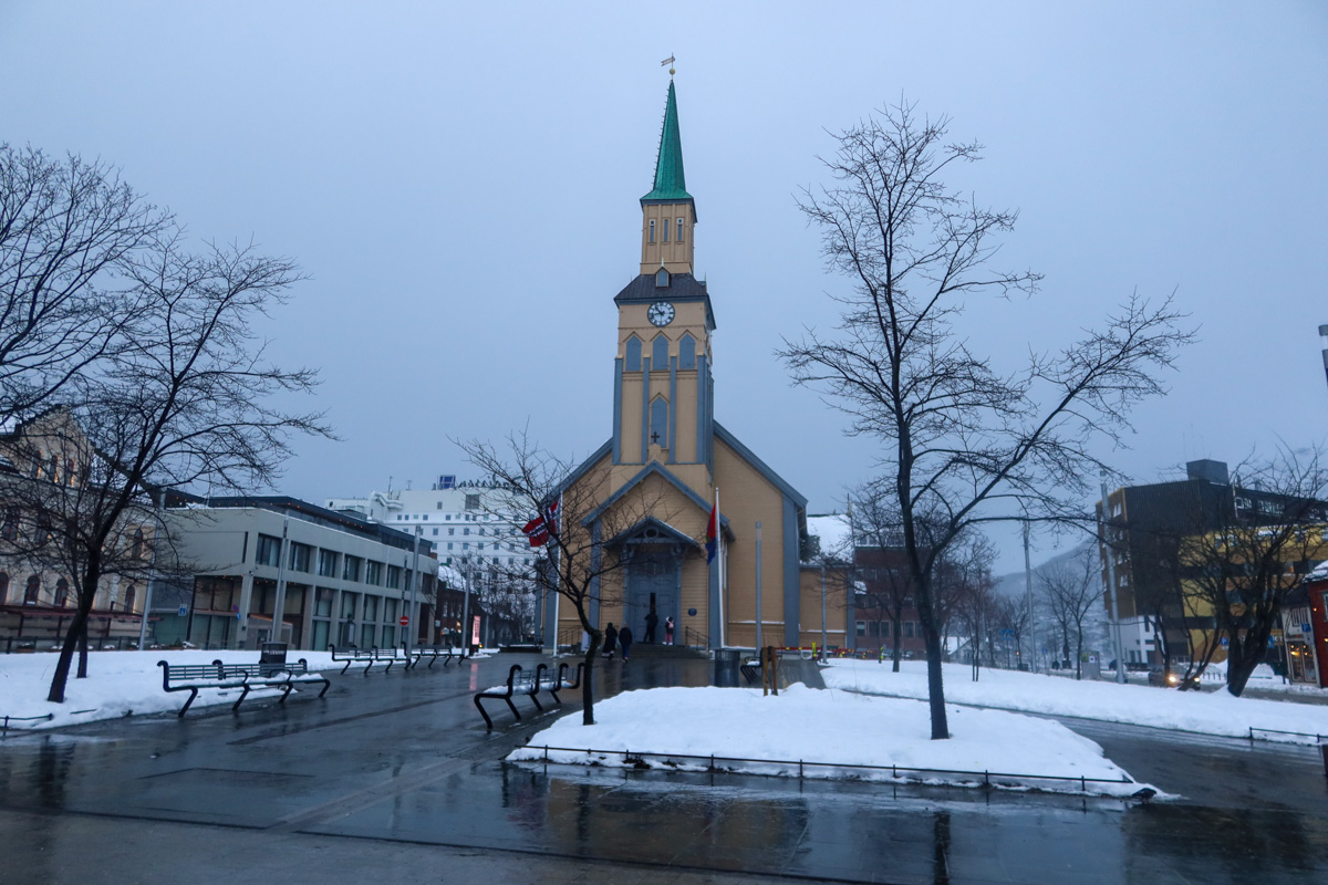 The yellow Tromso cathedral with a blue roof, with bare trees surrounding it, a cloudy sky and snow on the foreground