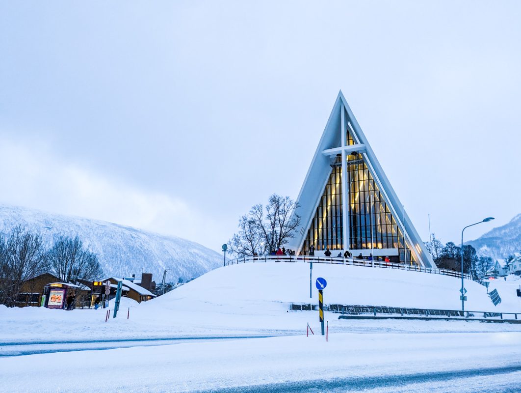 Arctic Cathedral, a modern building in the northern norway city of Tromso. The ground is covered in snow, the church is triangular in shape and there is a large cross in front.