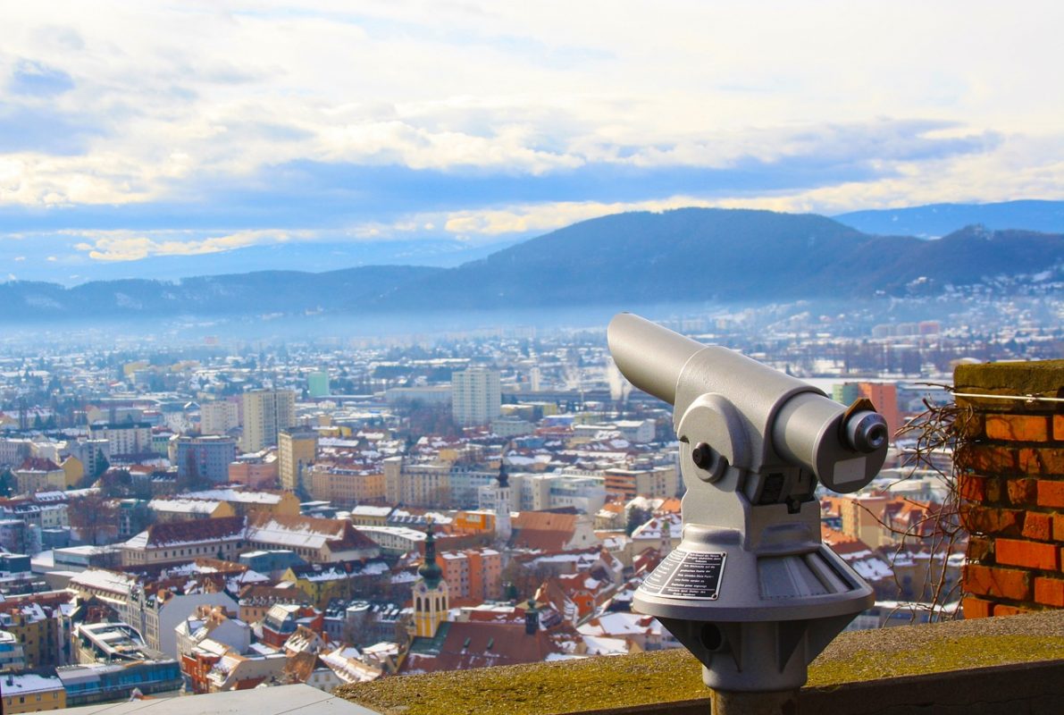 View of the city of Graz, which is the capital of the Styria region of Austria, with a telescope in the foreground and a range of houses int the background. 