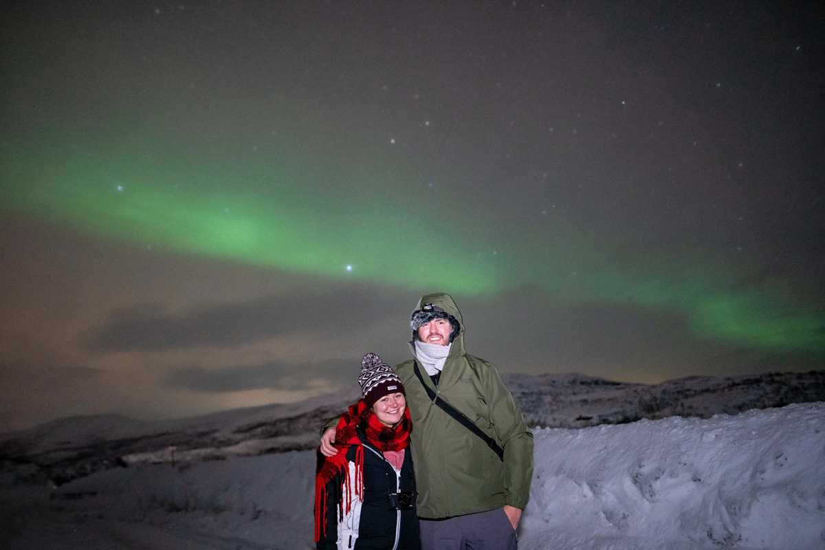 Woman and man standing infront of a dim glow of the northern lights near Tromso in Norway