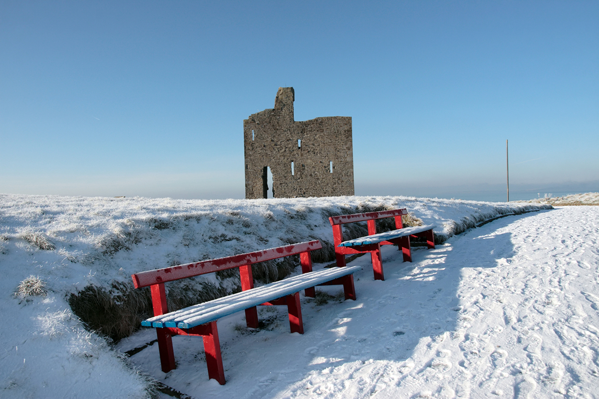 a seasonal snow covered view of atlantic ocean and ballybunion castle with red benches on a frosty snow covered winters day