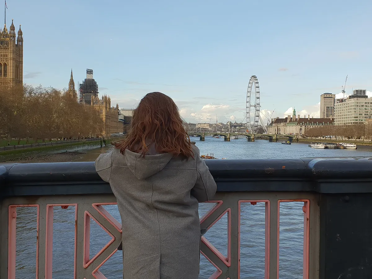 Girl looking over the River Thames and London Eye from a bridge. She is wearing a grey coat. The sky is blue.