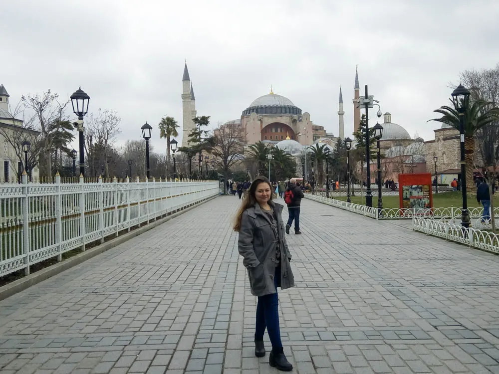 Girl standing infront of Hagia Sophia in Istanbul. The sky is grey and she is wearing trousers and a coat.