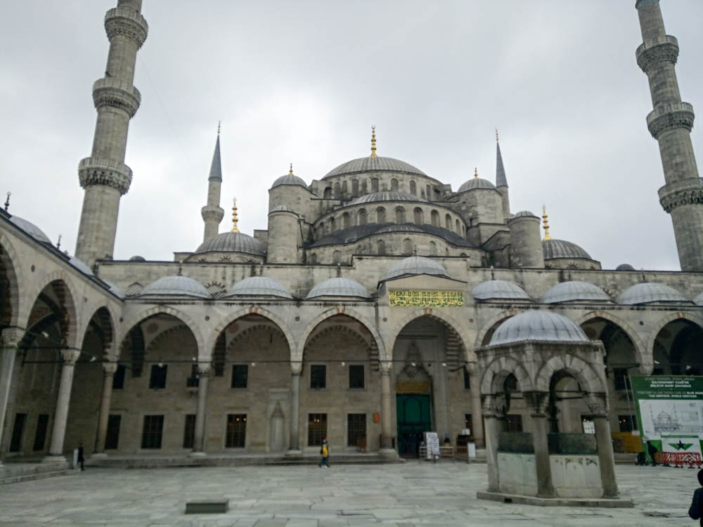 Inside the courtyard of the Blue mosque. Two turrets are on either side. The sky is cloudy.