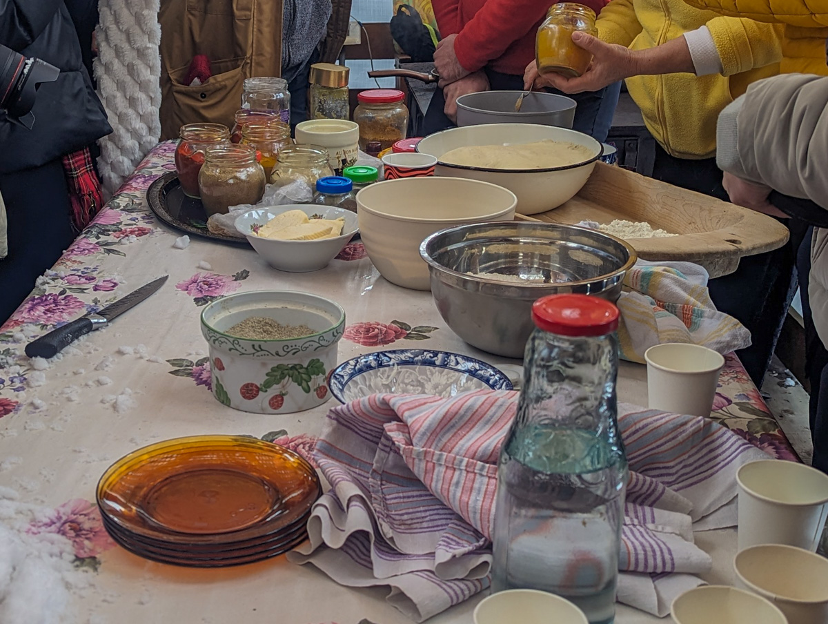 An outdoor cooking scene with bowls, jars of condiments, and a group of people preparing food on a table with a floral tablecloth.