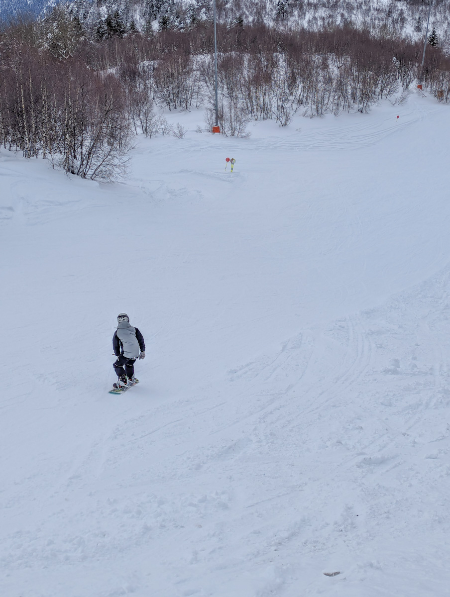 A snowboarder descending a snowy slope with ski lifts and sparse trees in the background in Mestia, Georgia.