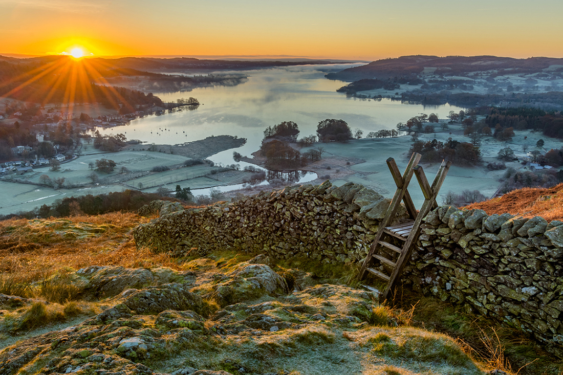 Wooden stile and stone wall with Lake Windermere in background with sun rising above horizon.