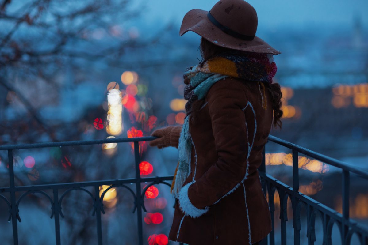 Seen from behind woman in brown hat and scarf outdoors in the city park in winter in sheepskin coat at night.