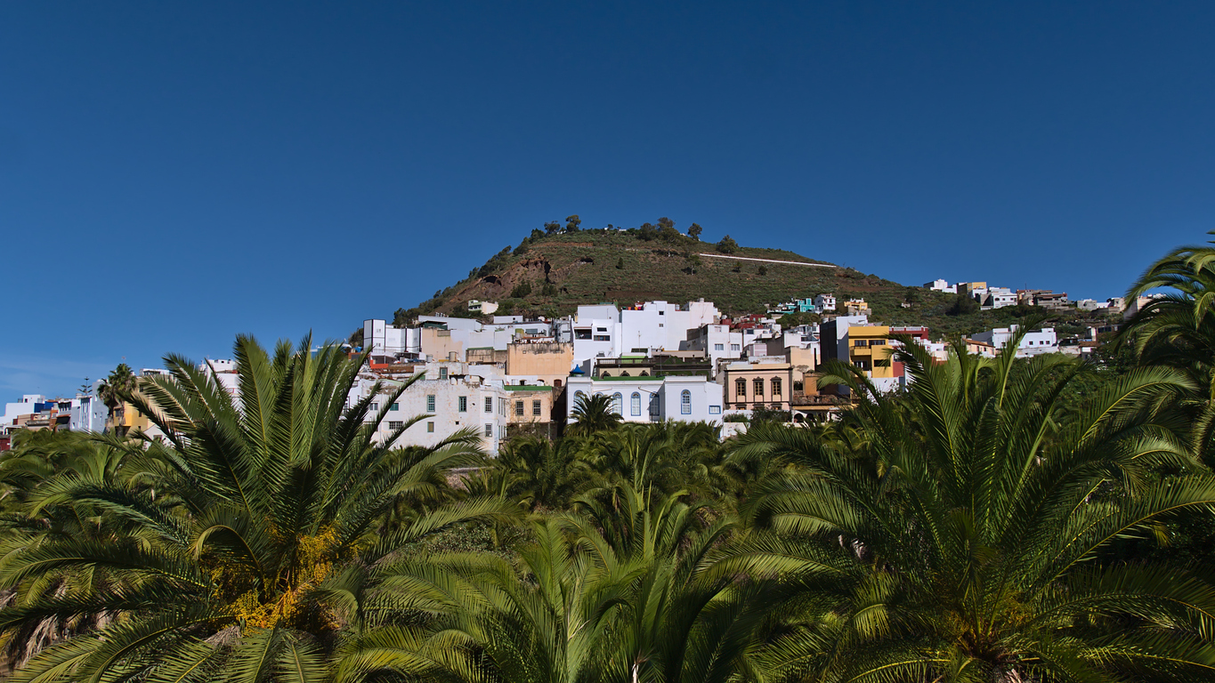 View of characteristic residential buildings in town Arucas in the north of Gran Canaria, Canary Islands, Spain on sunny day with hill and green palm trees in front in winter season.