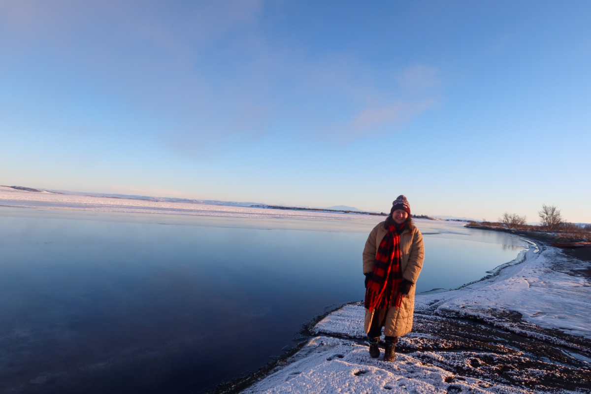 Blue thermal pool at the Golden Circle in winter, with bright blue in the left and a patch of snow. There's a girl in a beige coat standing in the snow by the lake.