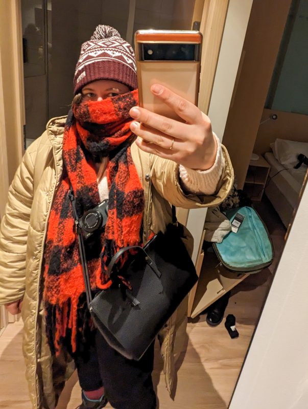 Girl taking a photo in a mirror wrapped up in a red and white scarf, maroon hat and beige coat.
