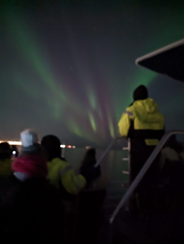 A crowd watching the Northern Lights on a boat trip just out of Reykjavik harbour, with green flashes in the sky.