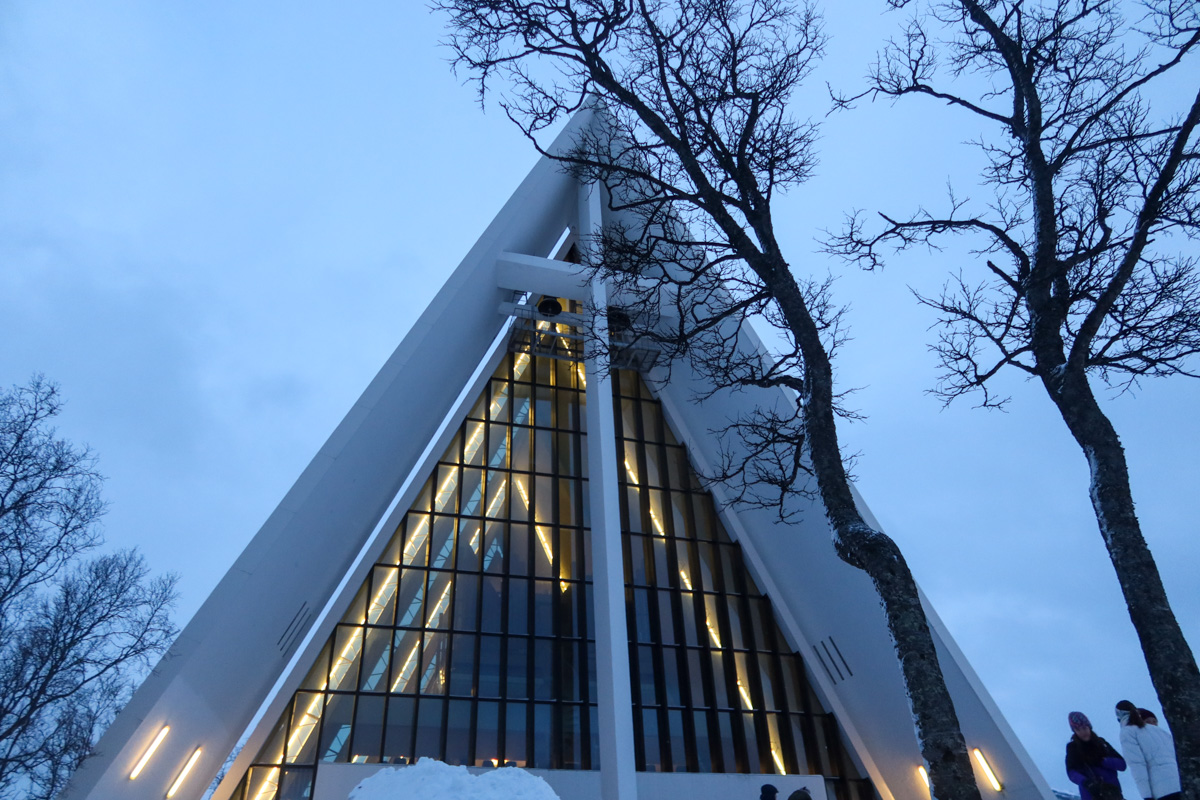 The roof of Arctic Cathedral, with a cross in the window, with lights in the background and bare trees in the foreground.