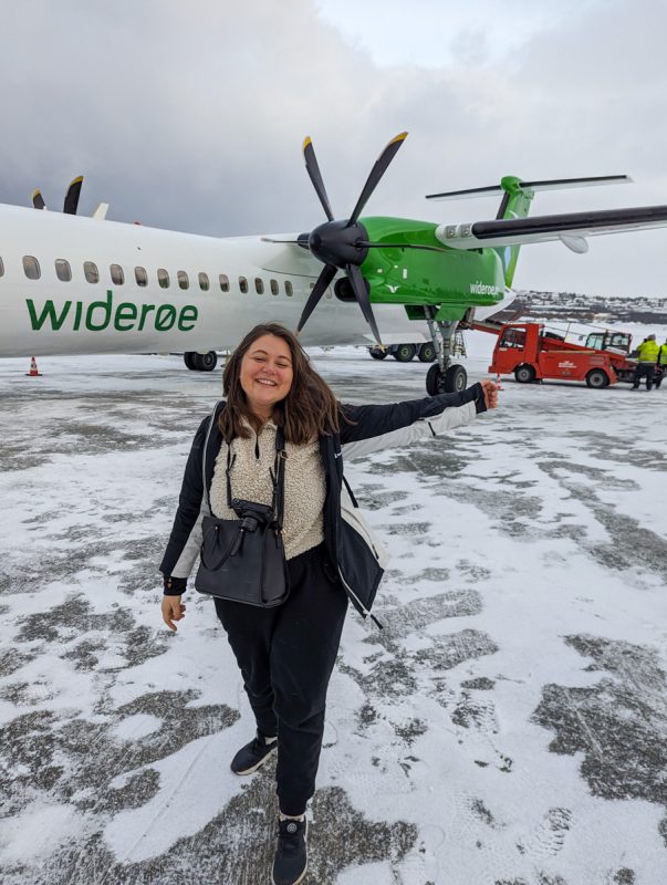 Girl standing in front of a Wideroe plane in Tromso, with snow on the ground. She is wearing black tracksuit bottoms, a cream jumper and black coat and has her arm in the air.