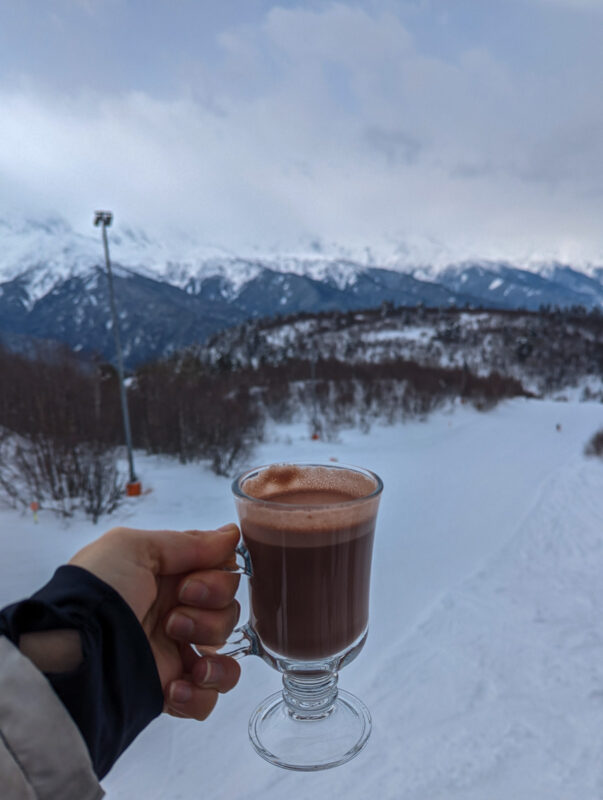 A hand holding a cup of hot chocolate with a scenic view of a snowy ski slope and mountain range in Mestia, Georgia.