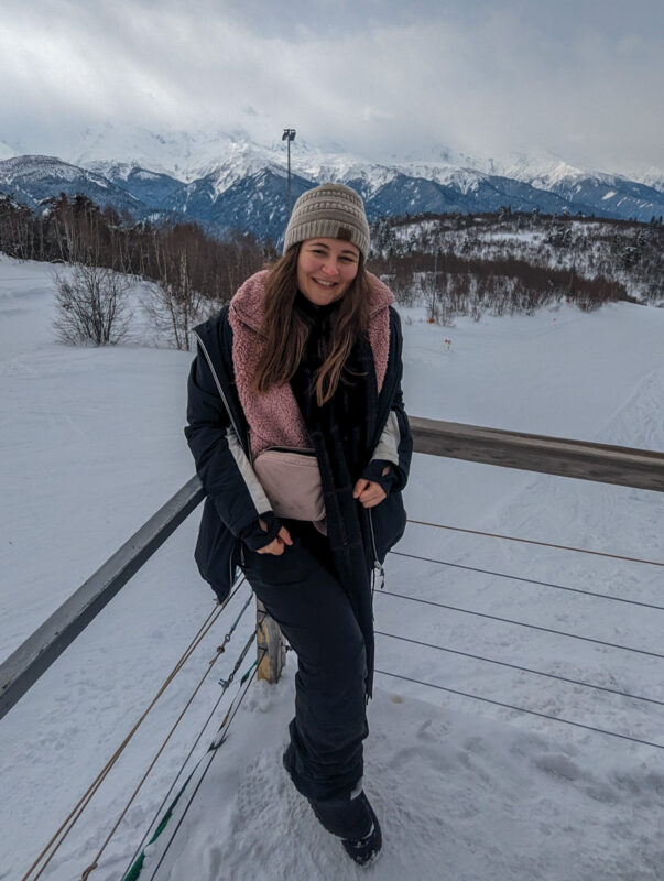 A smiling woman in winter clothing with a picturesque backdrop of snow-covered mountains in Mestia, Georgia.