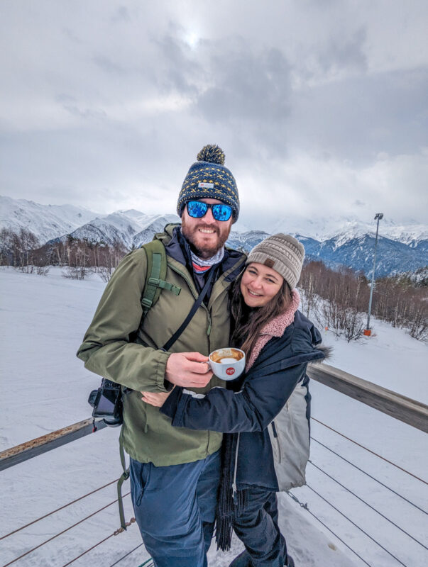 A happy couple holding a cup of coffee, standing on a snow-covered slope with a mountainous horizon in Mestia, Georgia.