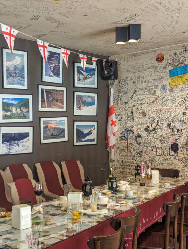 Interior of a cozy restaurant in Mestia, Georgia, with walls and ceiling covered in signatures and notes from visitors, Georgian flags, and pictures of mountainous landscapes.