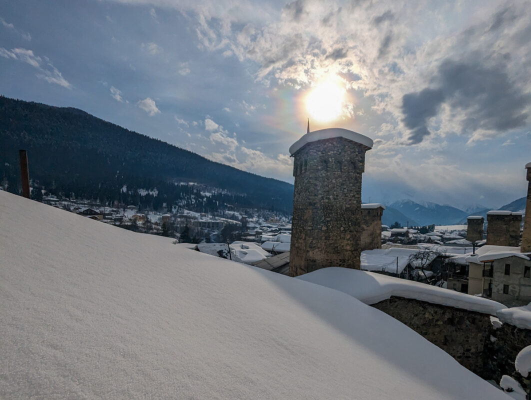 A snow-covered landscape at sunset with a traditional Svan tower and the sun peeking out from behind, casting a warm glow over Mestia, Georgia.