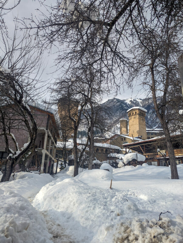 A picturesque view of a snow-blanketed street in Mestia, Georgia, with bare trees, traditional stone towers, and buildings against a backdrop of mountains
