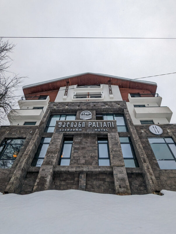 The facade of the Paliani Hotel in Mestia, Georgia, with a mix of stone and modern architecture, against a backdrop of a snowy landscape and a gray sky.